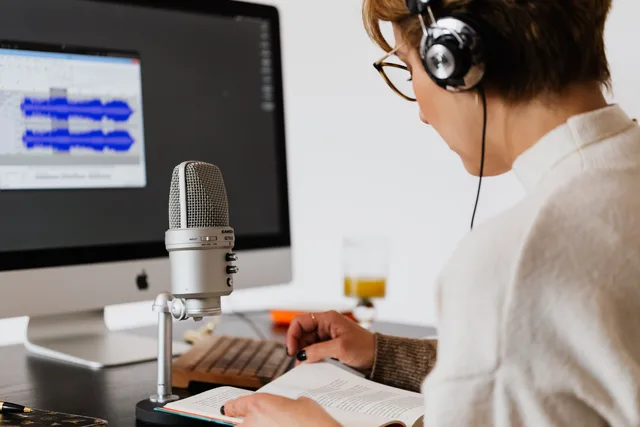 A woman using a microphone and computer to record and edit a podcast while reading from a book. Zielseiten für Audio-Werbung
